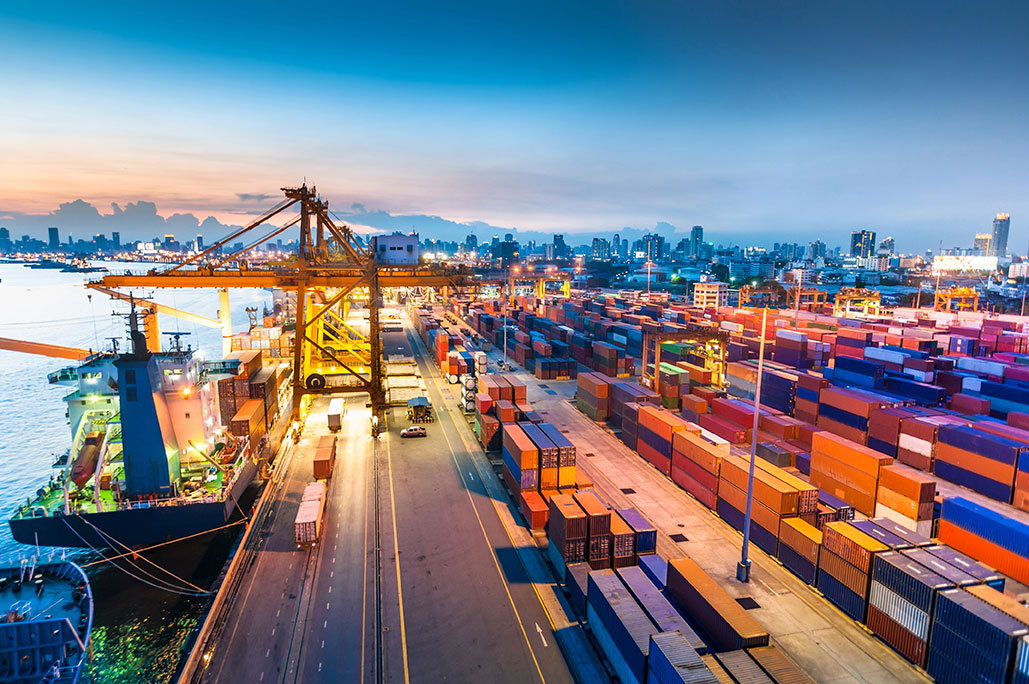 Shipping containers stacked at a port terminal with cranes loading cargo ships at dusk