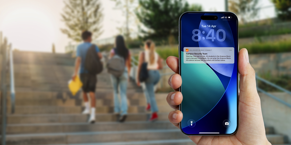 iPhone displaying an emergency broadcast notification for schools, with students walking on a school campus in the background